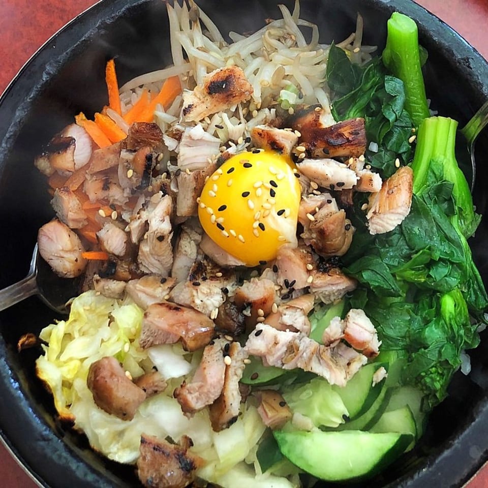 Bibimbap with grilled chicken, various vegetables, and a raw egg yolk in a black bowl.
